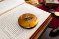 A round gold-toned Stratton compact mirror featuring an illustration of a ballerina in a black tutu, resting on an open book surrounded by vintage-style props.