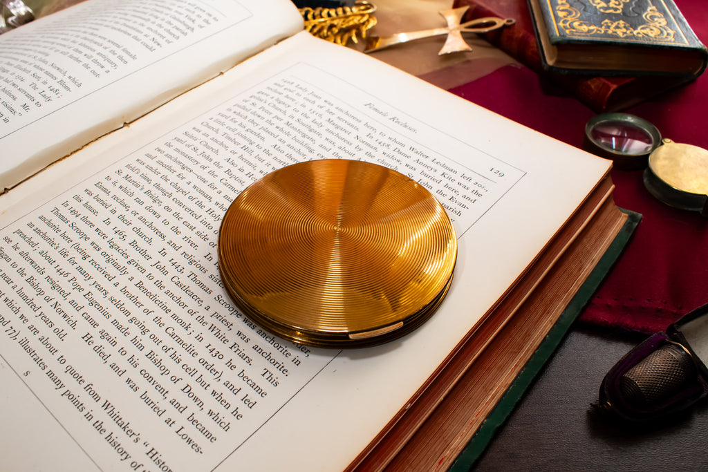 A round, polished gold Stratton powder compact  resting on an open book, surrounded by vintage books, a red cloth, and small accessories on a dark table.