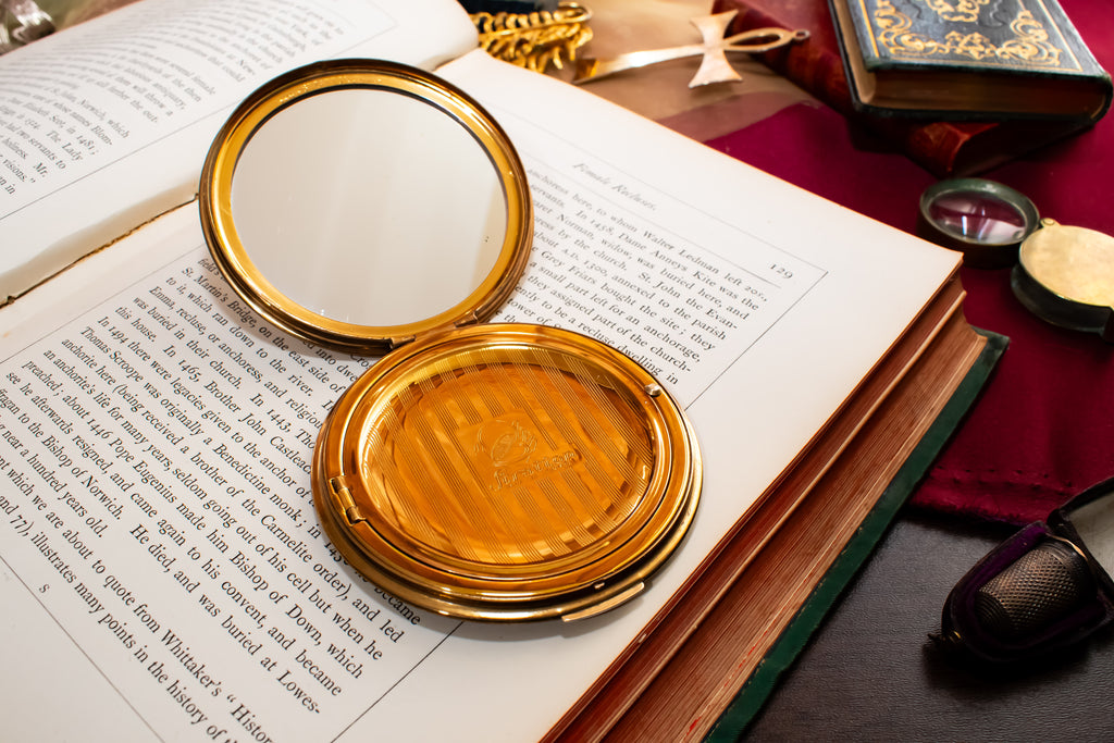 A round gold Stratton compact mirror with its lid open, resting on an open book surrounded by vintage-style props including old books, a red cloth, and a small round object on a dark tabletop.