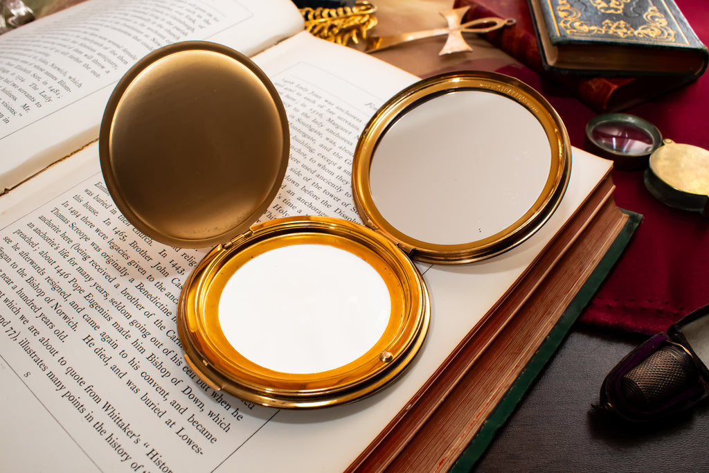 An open gold-framed Stratton compact mirror with a white interior, placed on top of an open book surrounded by vintage props including another book, a small box, and a dark surface.