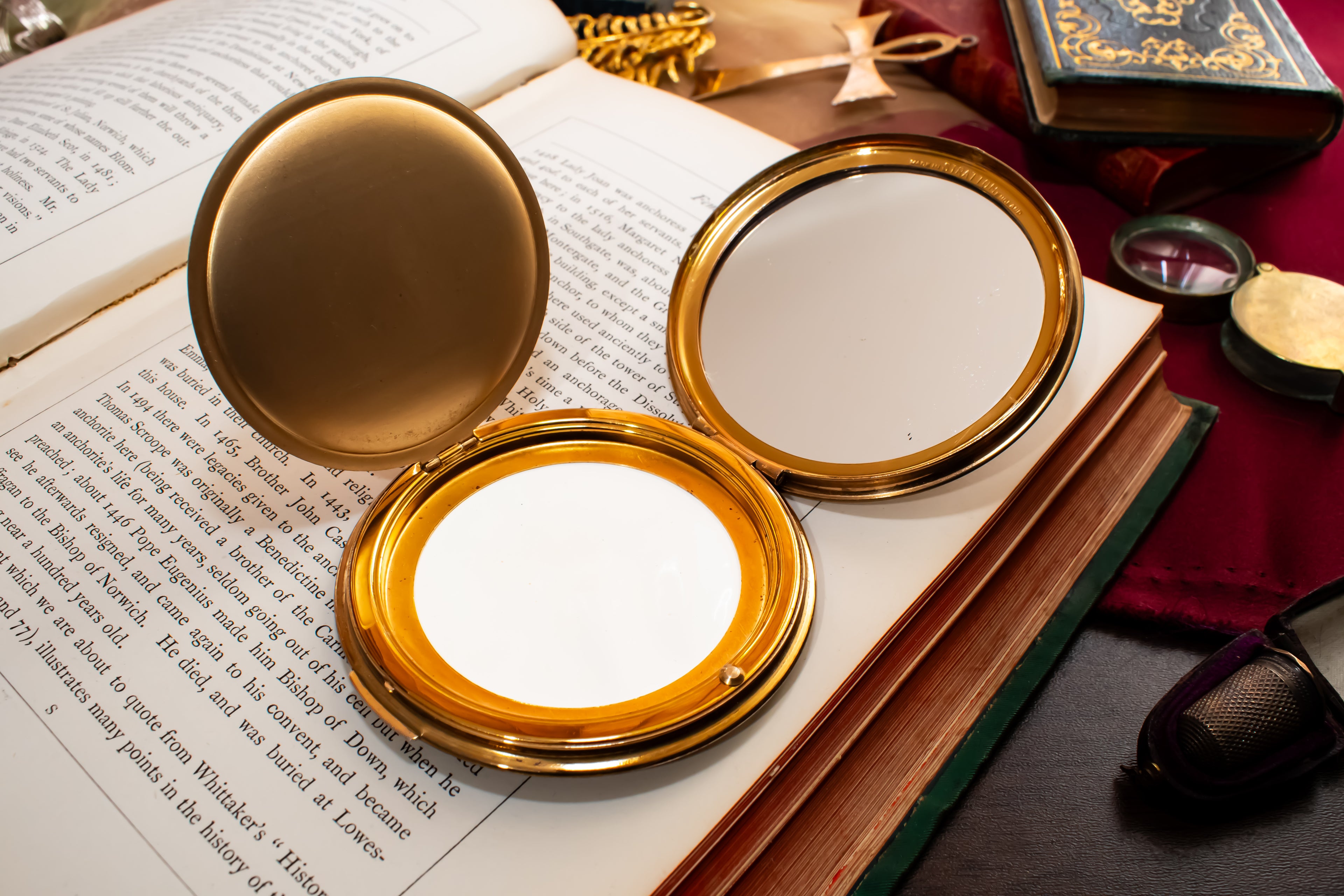 An open gold-framed Stratton compact mirror with a white interior, placed on top of an open book surrounded by vintage props including another book, a small box, and a dark surface.