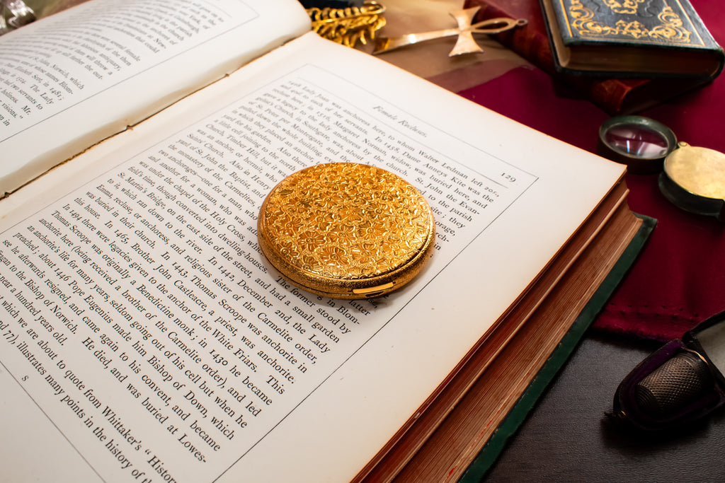 A round, textured gold Elisabeth Arden powder compact rests on an open book with printed text, surrounded by vintage books, a red cloth, and small accessories on a dark tabletop.