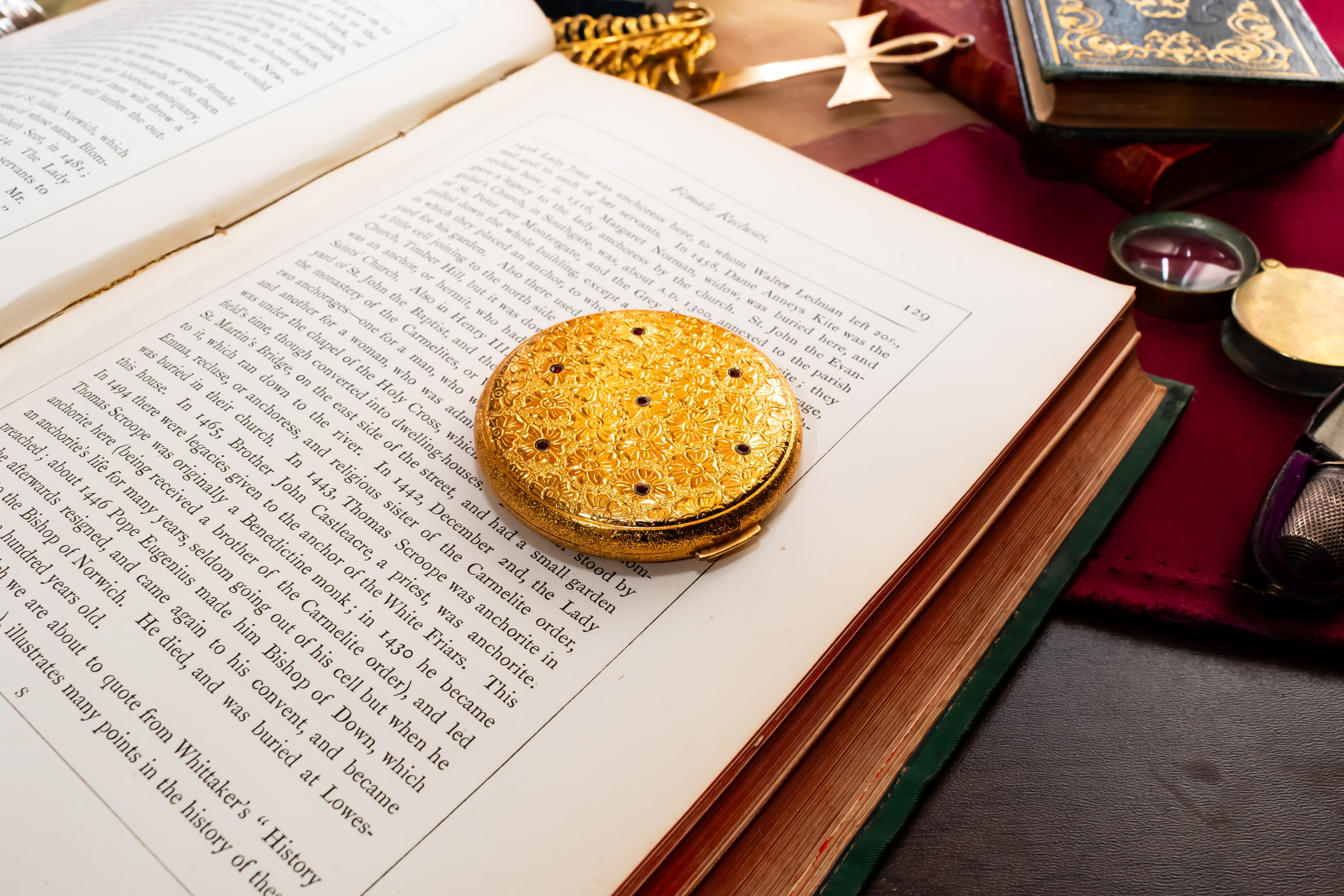 A round, textured gold-colored elisabeth Arden powder compact with small red garnet cabochons resting on an open book with printed text. The book is styled on a dark wooden table with other vintage books, a red cloth, and reading accessories in the background.
