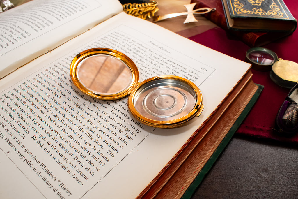 A round gold-toned Elisabeth Arden powder compact with an open lid showing a clear mirror and a silver-toned inner plate, resting on an open book surrounded by vintage books and mystical props.