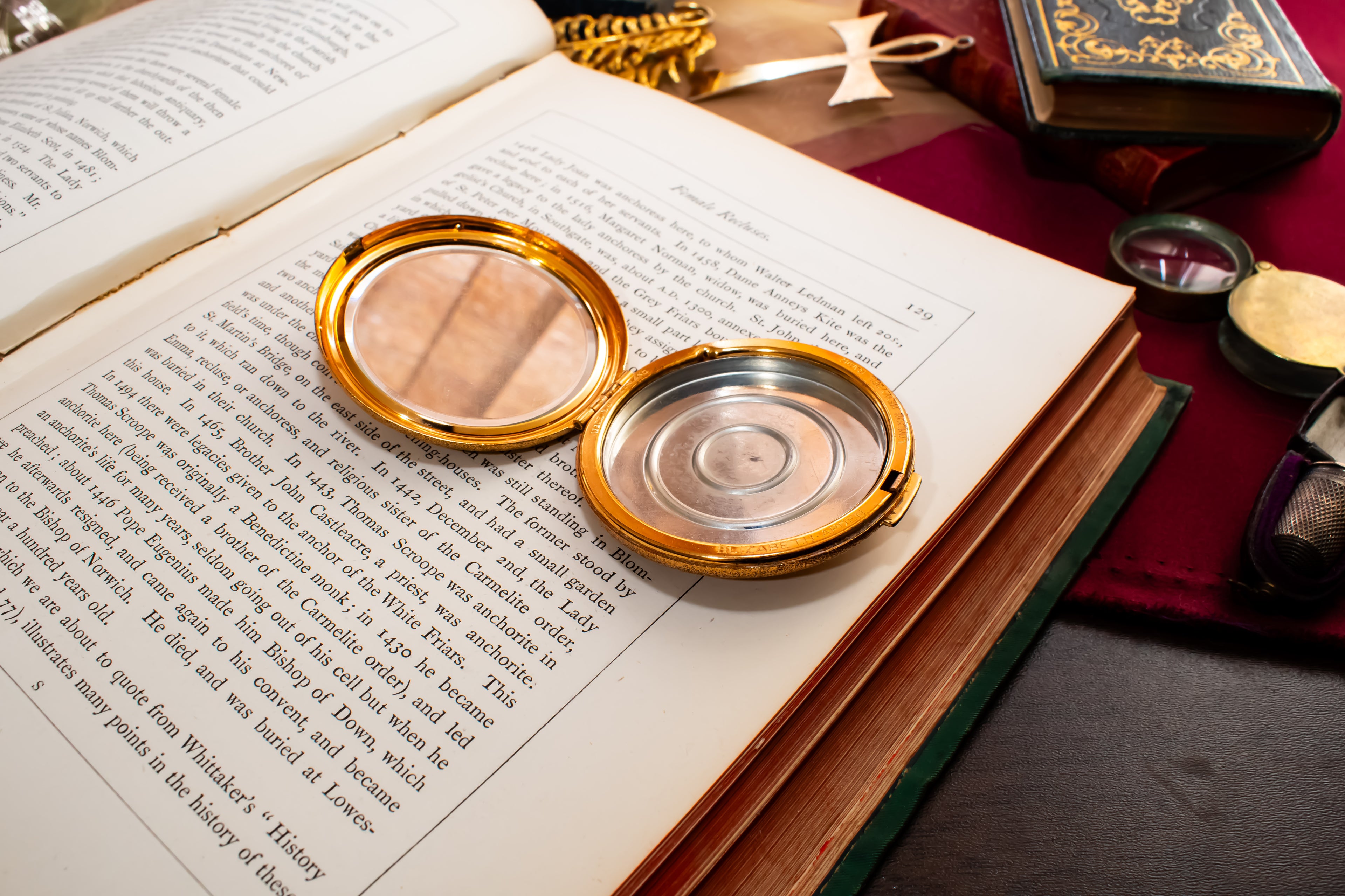 A round gold-toned Elisabeth Arden powder compact with an open lid showing a clear mirror and a silver-toned inner plate, resting on an open book surrounded by vintage books and mystical props.
