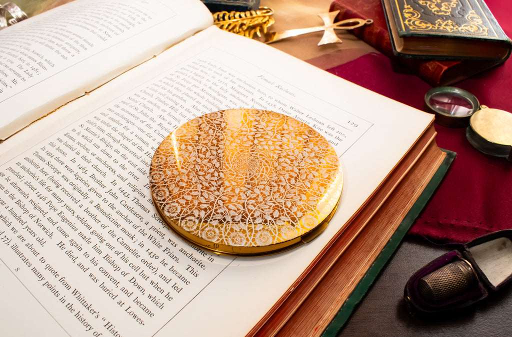 An ornate round gold and white filigree powder compact with intricate lace-like detailing, displayed on the pages of an open book surrounded by vintage books and reading accessories.