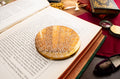 An ornate round gold and white filigree powder compact with intricate lace-like detailing, displayed on the pages of an open book surrounded by vintage books and reading accessories.