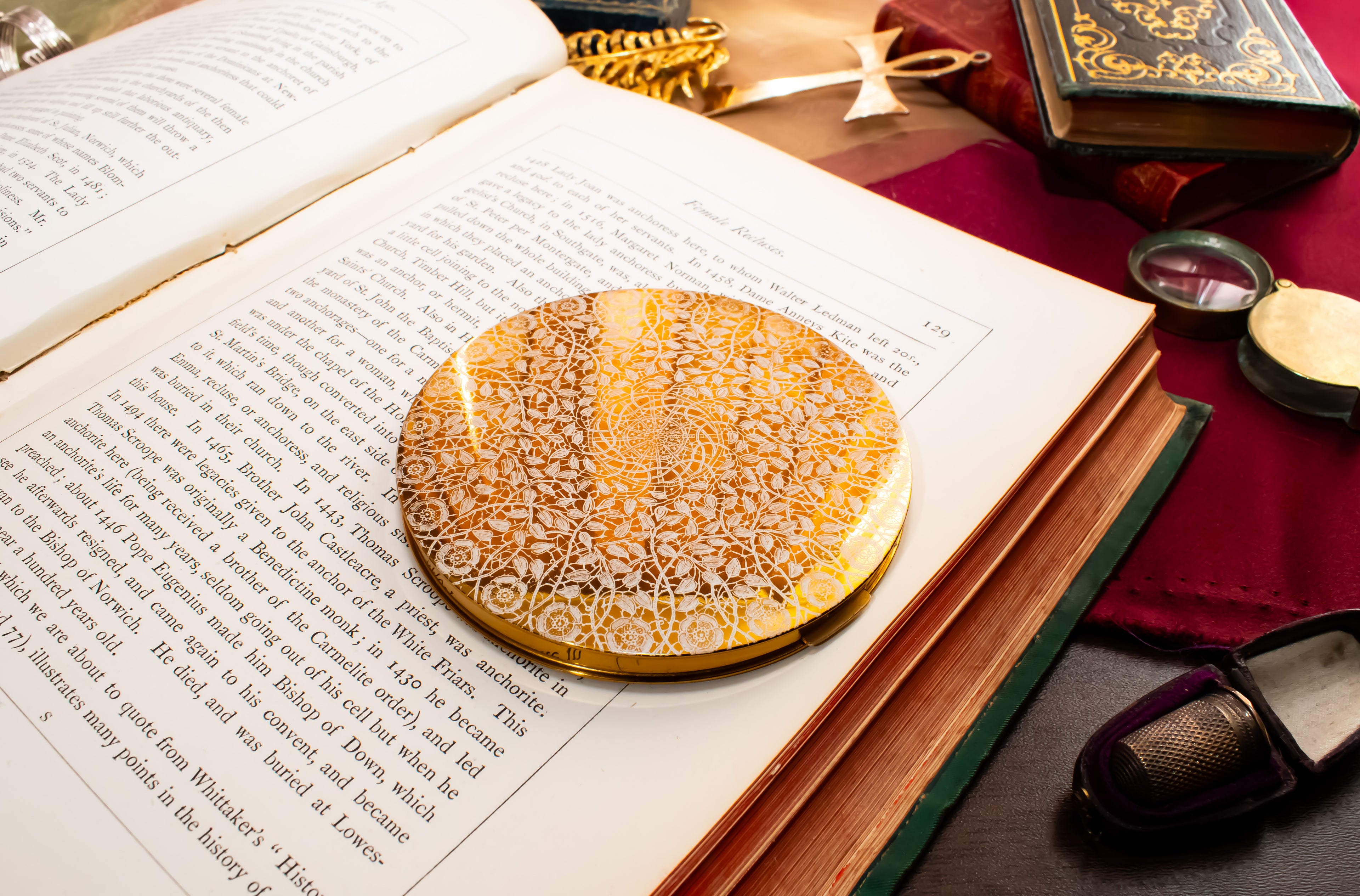 An ornate round gold and white filigree powder compact with intricate lace-like detailing, displayed on the pages of an open book surrounded by vintage books and reading accessories.
