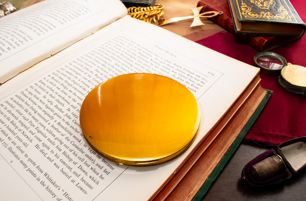 A round, polished gold compact mirror resting on the pages of an open book, surrounded by vintage books, a red cloth, and small accessories on a dark wooden surface.