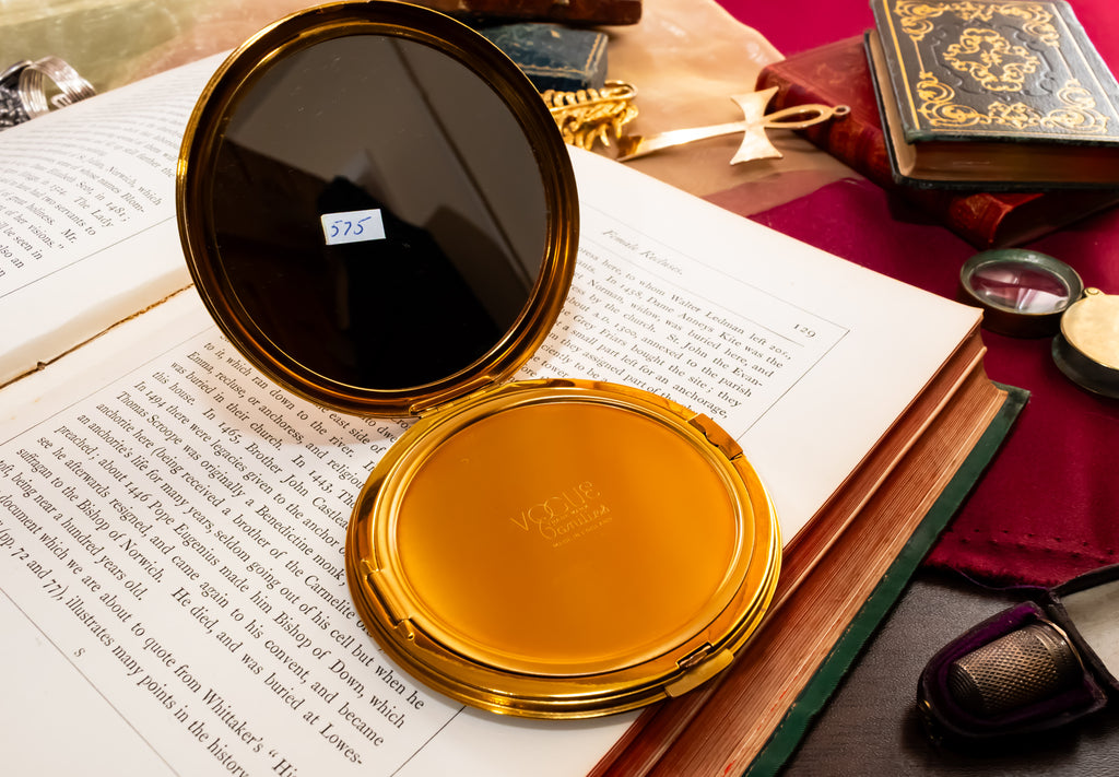 An open gold-toned compact mirror with a mirror interior, placed on top of an open book surrounded by vintage books, a red cloth, and small accessories.