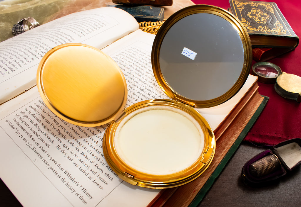 An open round gold compact mirror resting on an open book, with a small white powder pan visible inside. Other vintage-style objects and books are arranged around it on a dark surface.