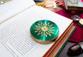 A round Margaret Rose green and gold compact mirror with a floral sunburst design, resting on the pages of an open book surrounded by vintage props.