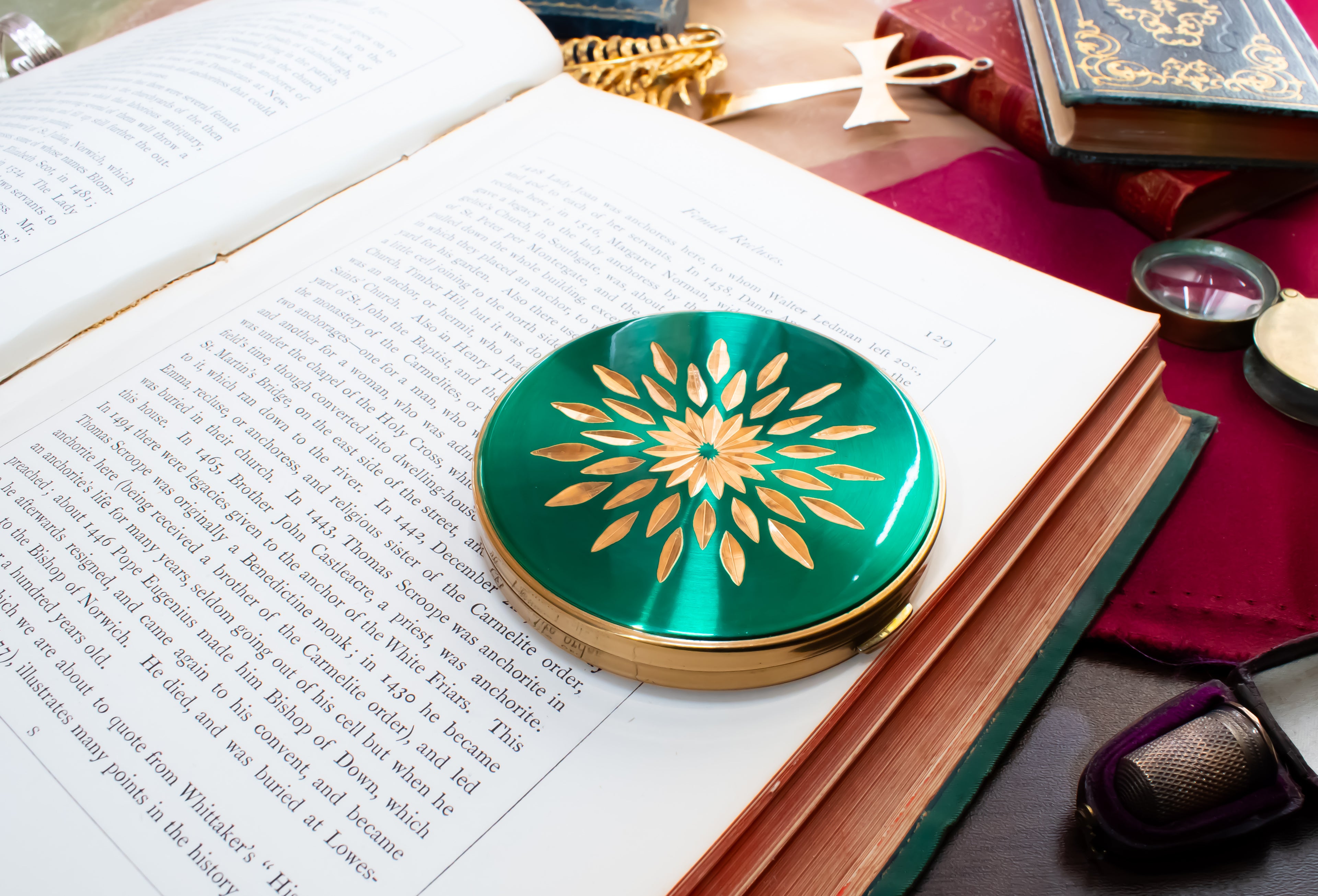 A round Margaret Rose green and gold compact mirror with a floral sunburst design, resting on the pages of an open book surrounded by vintage props.