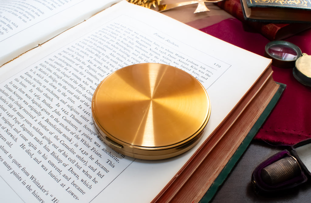 A round, brushed gold metal Margaret Rose powder compact resting on an open book, surrounded by vintage-style reading props including a magnifying glass, glasses, and another book on a dark red cloth.
