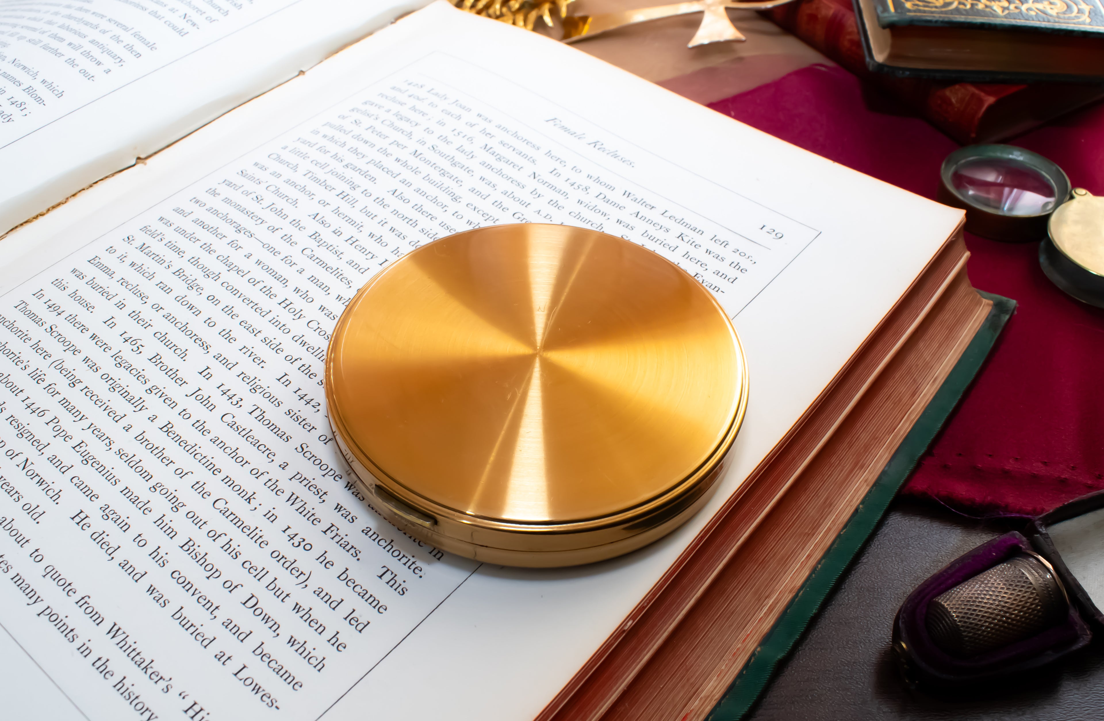 A round, brushed gold metal Margaret Rose powder compact resting on an open book, surrounded by vintage-style reading props including a magnifying glass, glasses, and another book on a dark red cloth.
