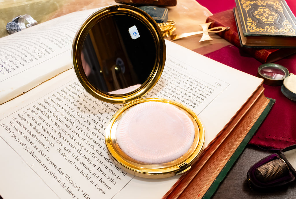 A round gold  Margaret Rose compact mirror with a white cushion foundation puff, displayed open on top of an open book surrounded by vintage-style props.