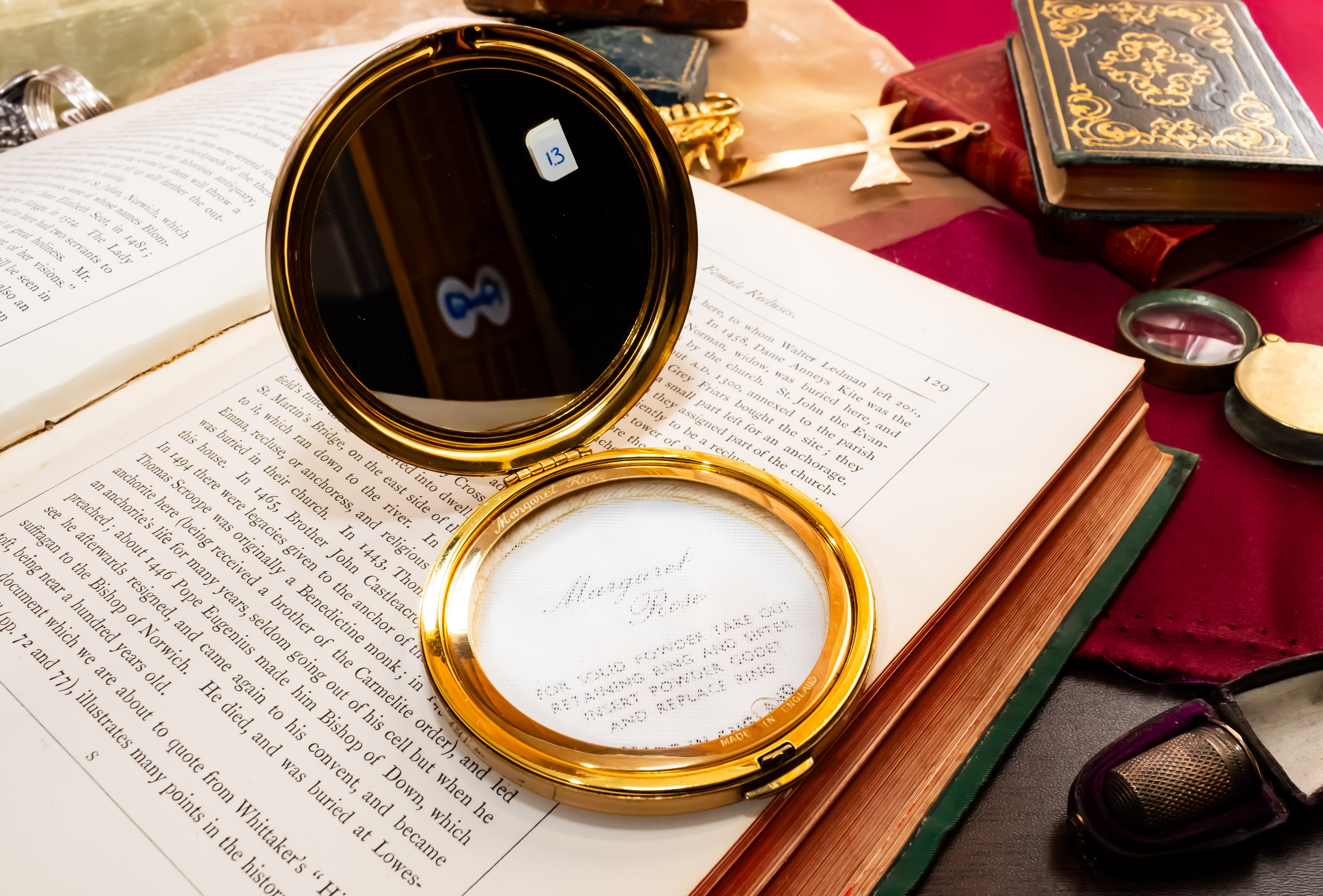 A gold-framed vintage Margaret Rose compact mirror with a white surface labeled 'Margaret Rose' is displayed open on top of an open book, surrounded by old books, a red cloth, and other antique-style props.