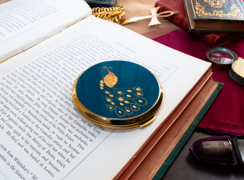A round Stratton compact mirror with a gold-trimmed lid featuring a blue peacock illustration, resting on an open book surrounded by vintage props.