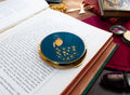 A round Stratton compact mirror with a gold-trimmed lid featuring a blue peacock illustration, resting on an open book surrounded by vintage props.