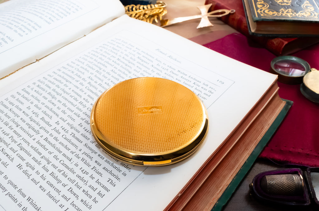 A round gold Stratton compact case with a textured metallic finish and a small embossed emblem, resting on the pages of an open book surrounded by vintage study props.
