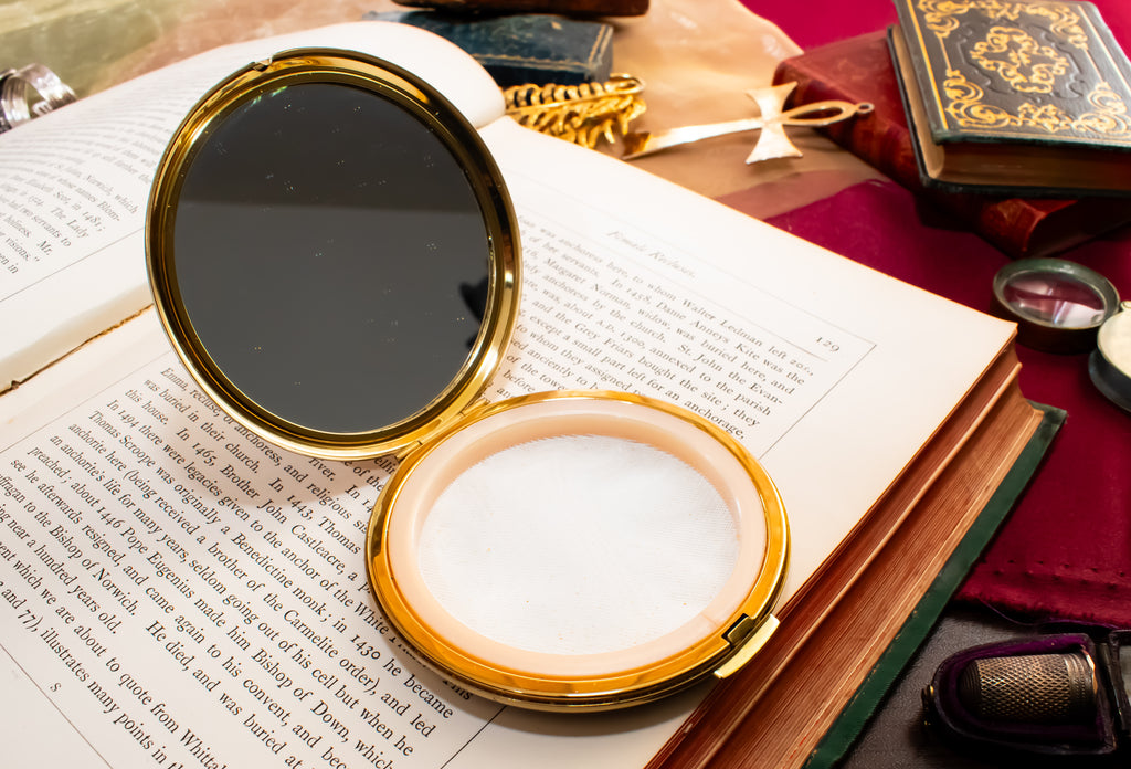 An open round Stratton gold compact mirror with a white interior sits on an open book surrounded by vintage books, a small magnifying glass, and a purple object on a red cloth background.