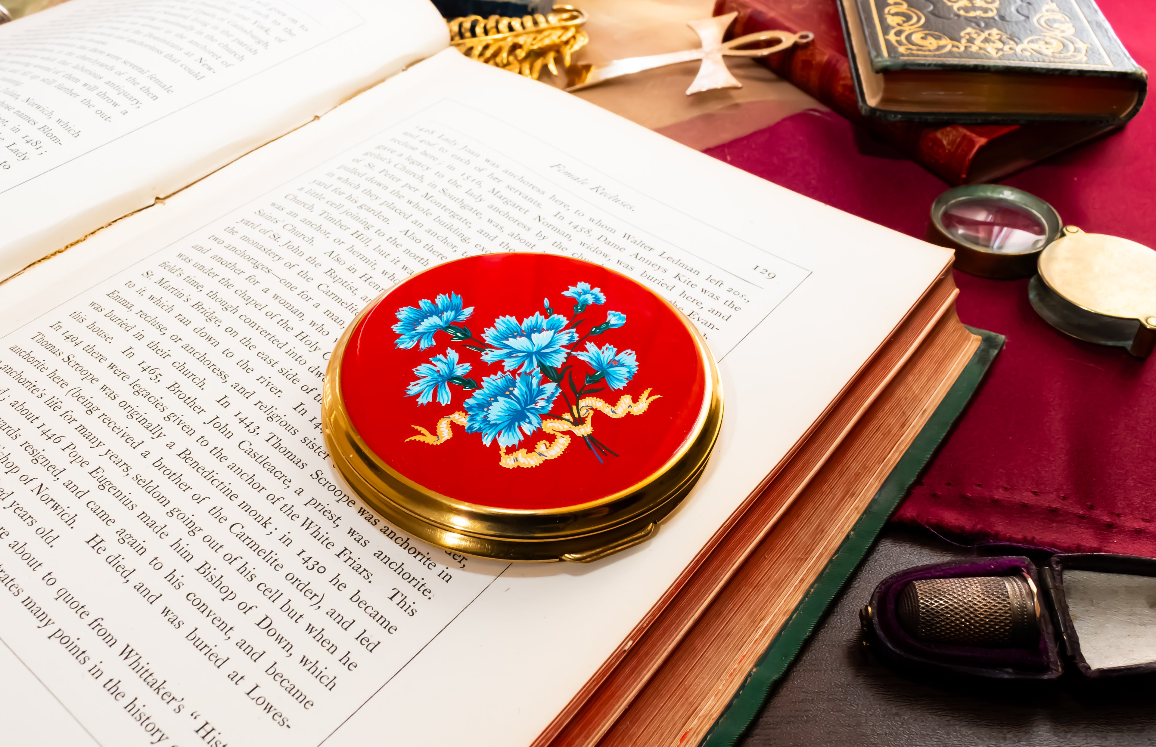 A round red Mascot powder compact with gold trim and blue floral motif, resting on an open book surrounded by vintage props including old books, a magnifying glass, and a miniature camera.