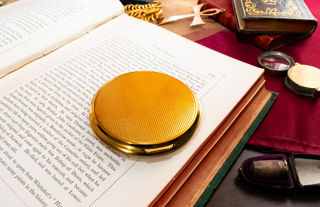 A round gold Mascot compact mirror with a textured metallic surface, resting on the pages of an open book. The scene is styled with vintage props including a red cloth, a vintage typewriter, and old books.