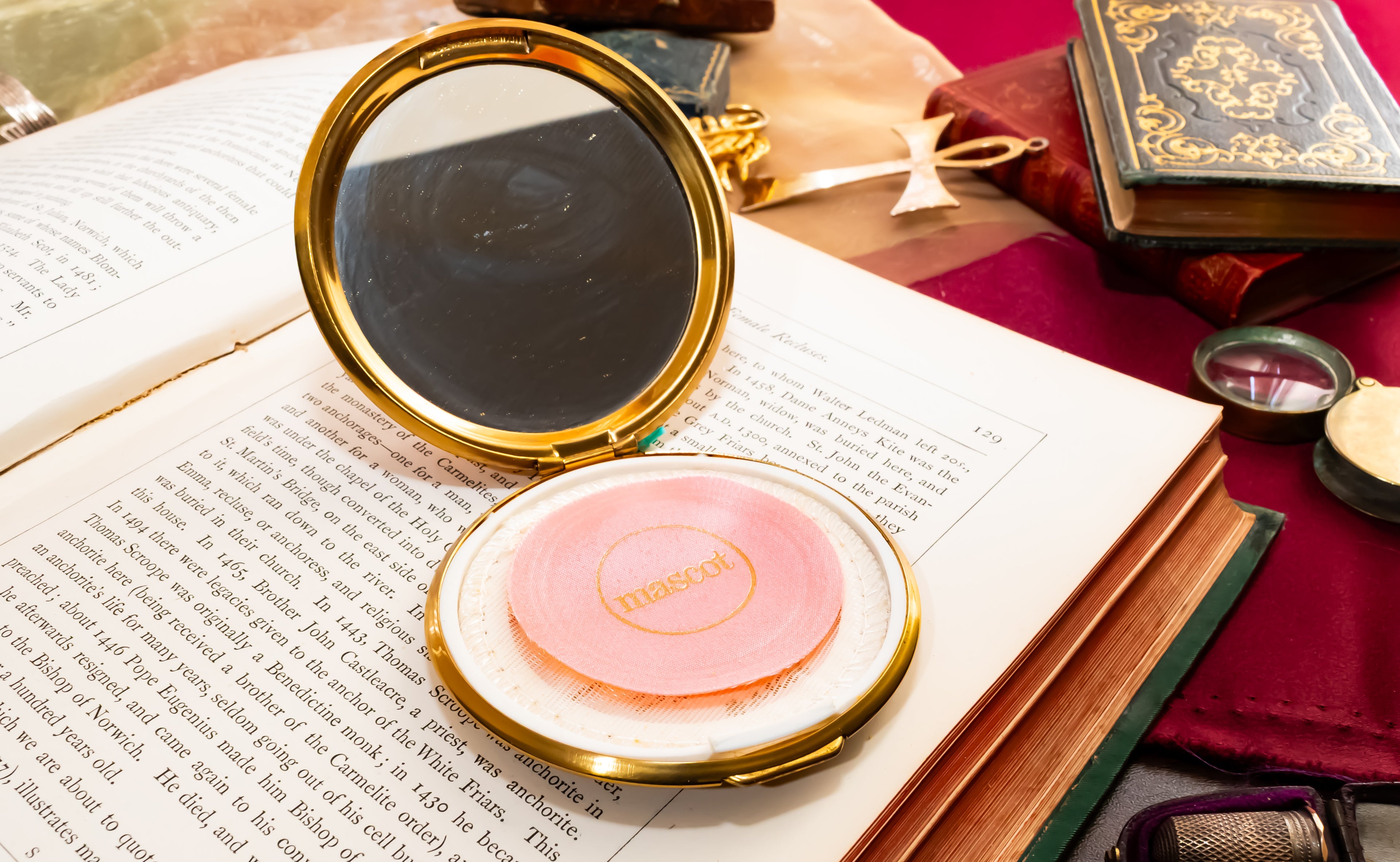 A round compact mirror with a gold frame and pink pressed powder sits open on an open book, surrounded by vintage-style props including old books, a red cloth, and small accessories.