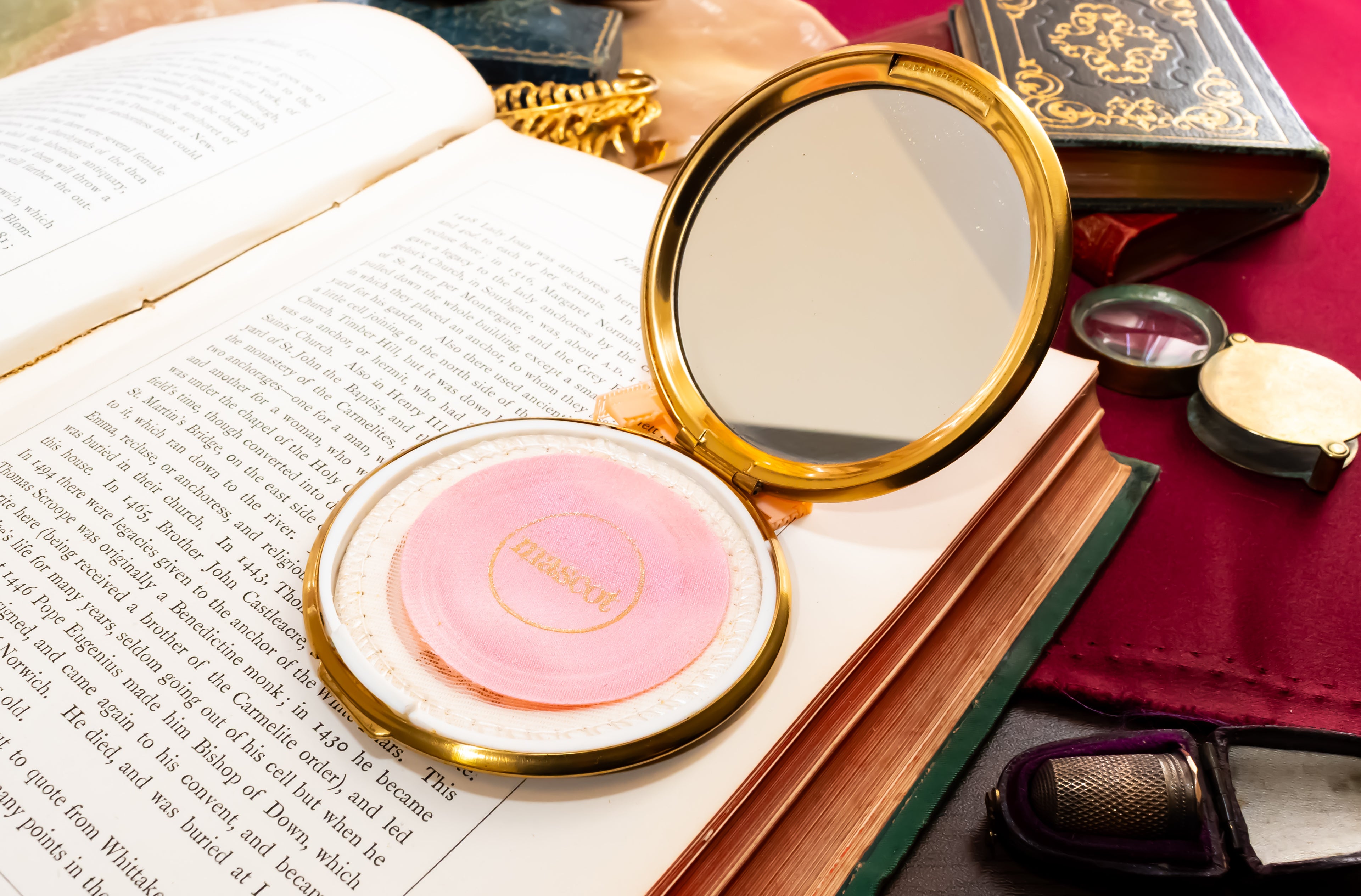 A round compact mirror with a gold frame and a pink pressed powder insert labeled 'Mascot', resting on an open book surrounded by vintage-style props including books, a red fabric surface, and small accessories.