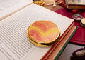 A round gold-plated coty powder compact with a red and yellow wavy patterned lens rests on the pages of an open book, surrounded by vintage books and reading accessories.