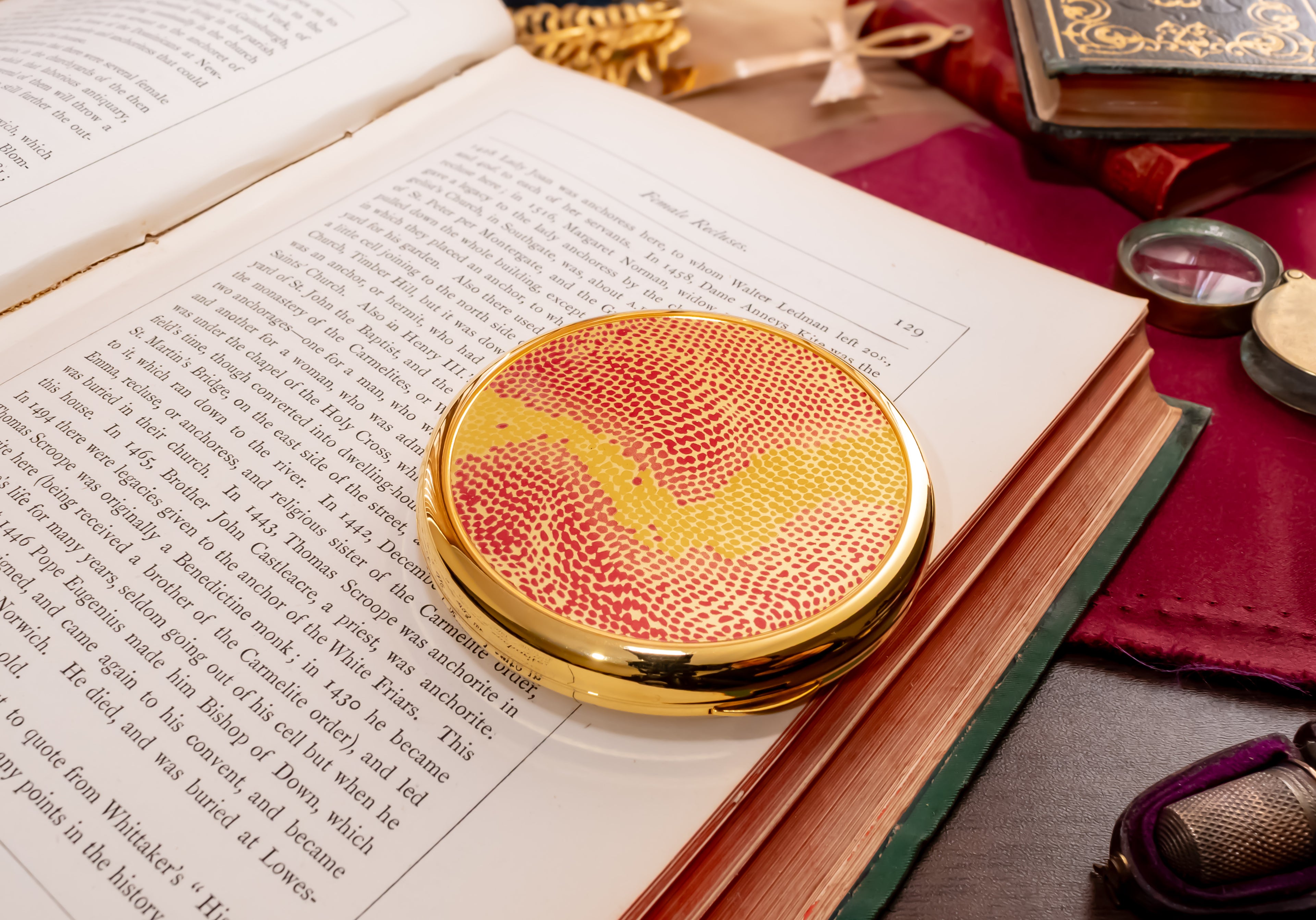 A round gold-plated coty powder compact with a red and yellow wavy patterned lens rests on the pages of an open book, surrounded by vintage books and reading accessories.