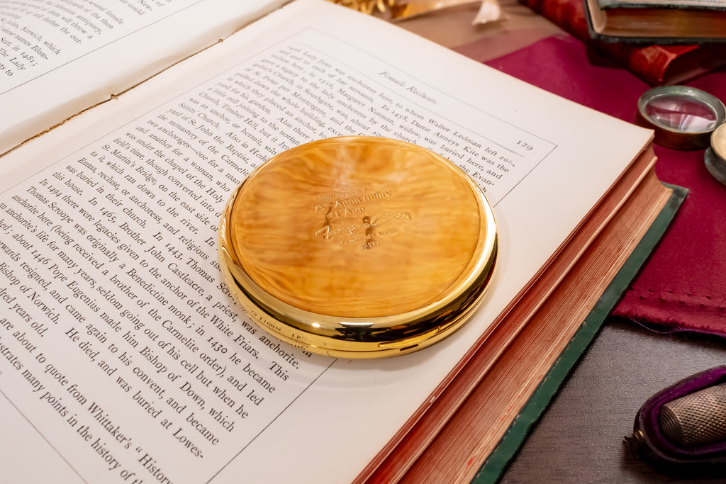 A round, polished gold colored coty compact, resting on the pages of an open book. The background features a vintage desk setup with a purple leather pad, a magnifying glass, and other antique items.