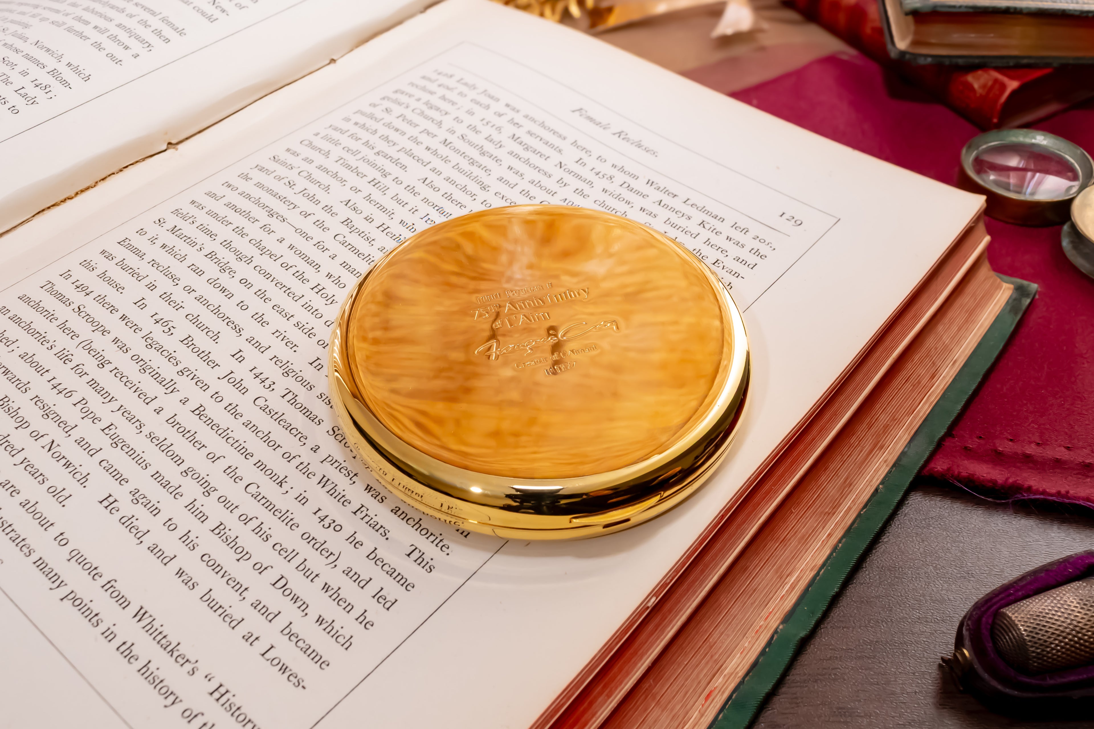 A round, polished gold colored coty compact, resting on the pages of an open book. The background features a vintage desk setup with a purple leather pad, a magnifying glass, and other antique items.
