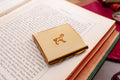 A square gold-toned compact case with a small dog silhouette emblem on top, resting on an open book.