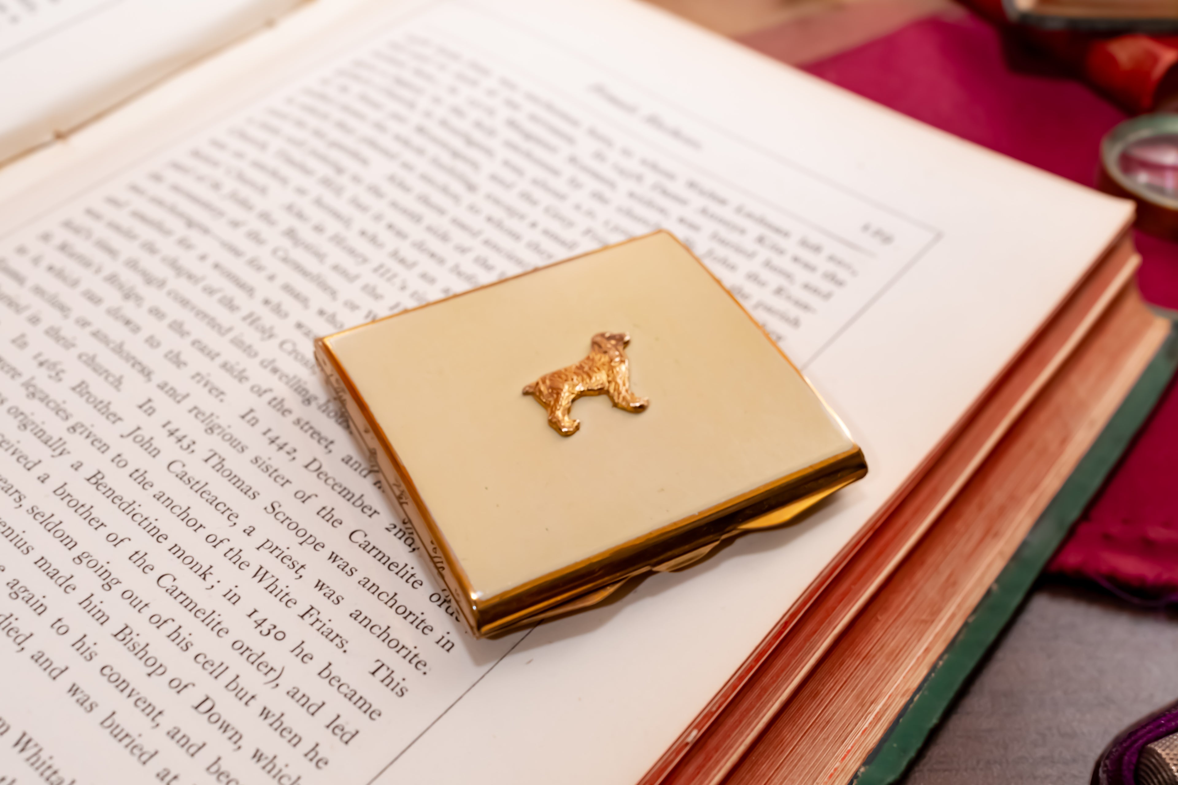 A square gold-toned compact case with a small dog silhouette emblem on top, resting on an open book.