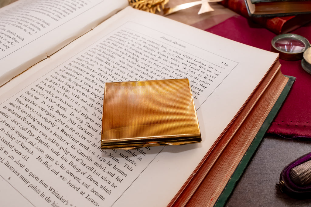 A square gold metal Stratton powder compact with a textured, brushed finish resting on an open book. The book is surrounded by vintage props including a purple leather-bound book, a small brass magnifying glass, and other antique items on a dark wooden table.