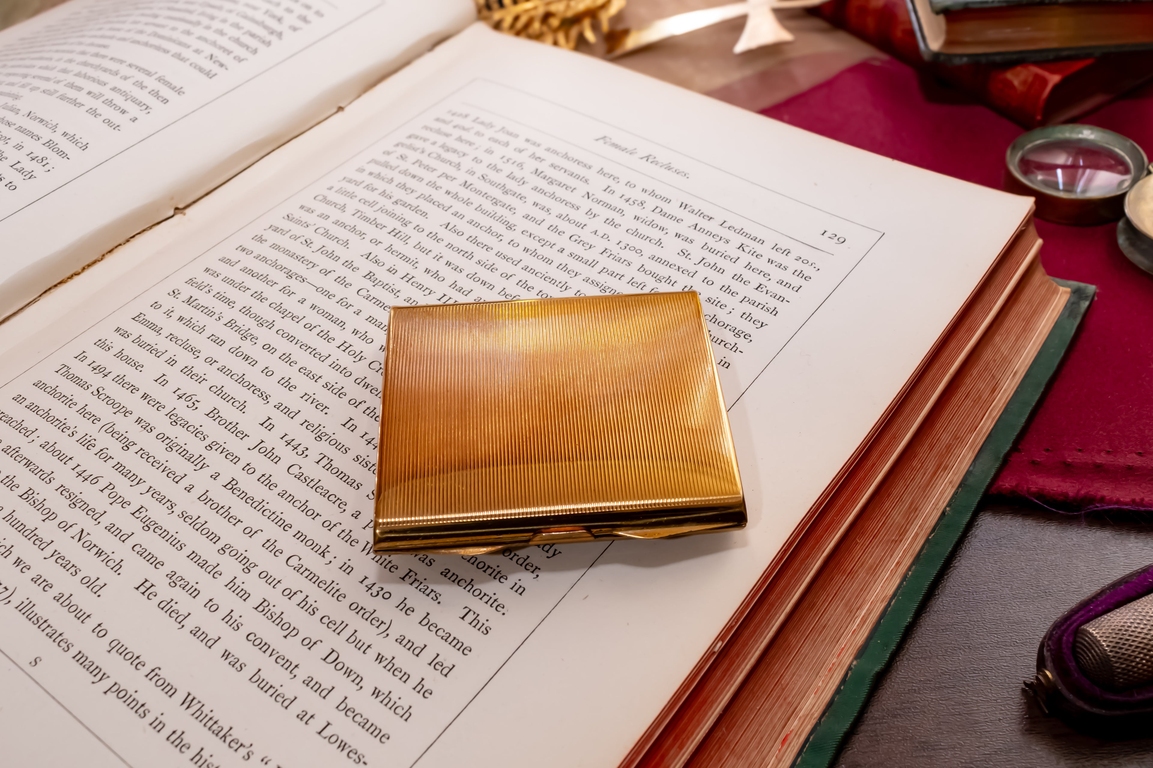 A square gold metal Stratton powder compact with a textured, brushed finish resting on an open book. The book is surrounded by vintage props including a purple leather-bound book, a small brass magnifying glass, and other antique items on a dark wooden table.