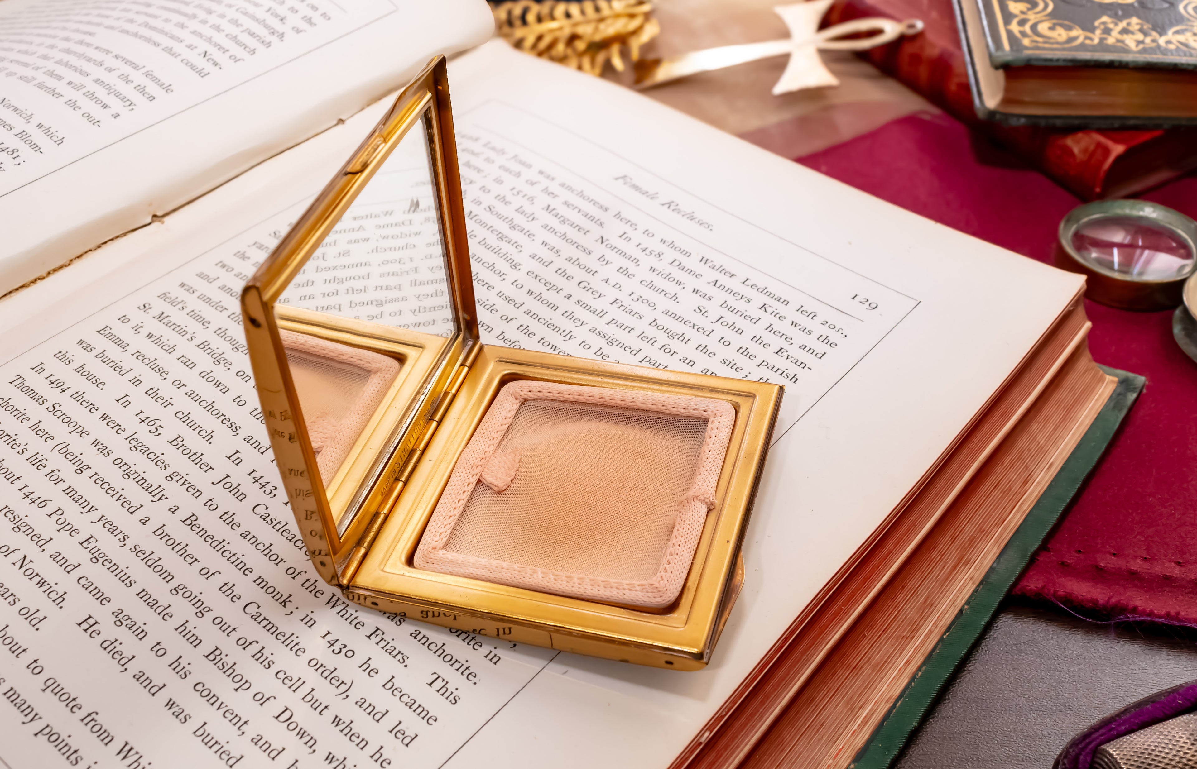 An open Stratton gold compact mirror with a light beige pressed powder inside, placed on top of an open book surrounded by vintage-style props including old books, a magnifying glass, and a ribbon.