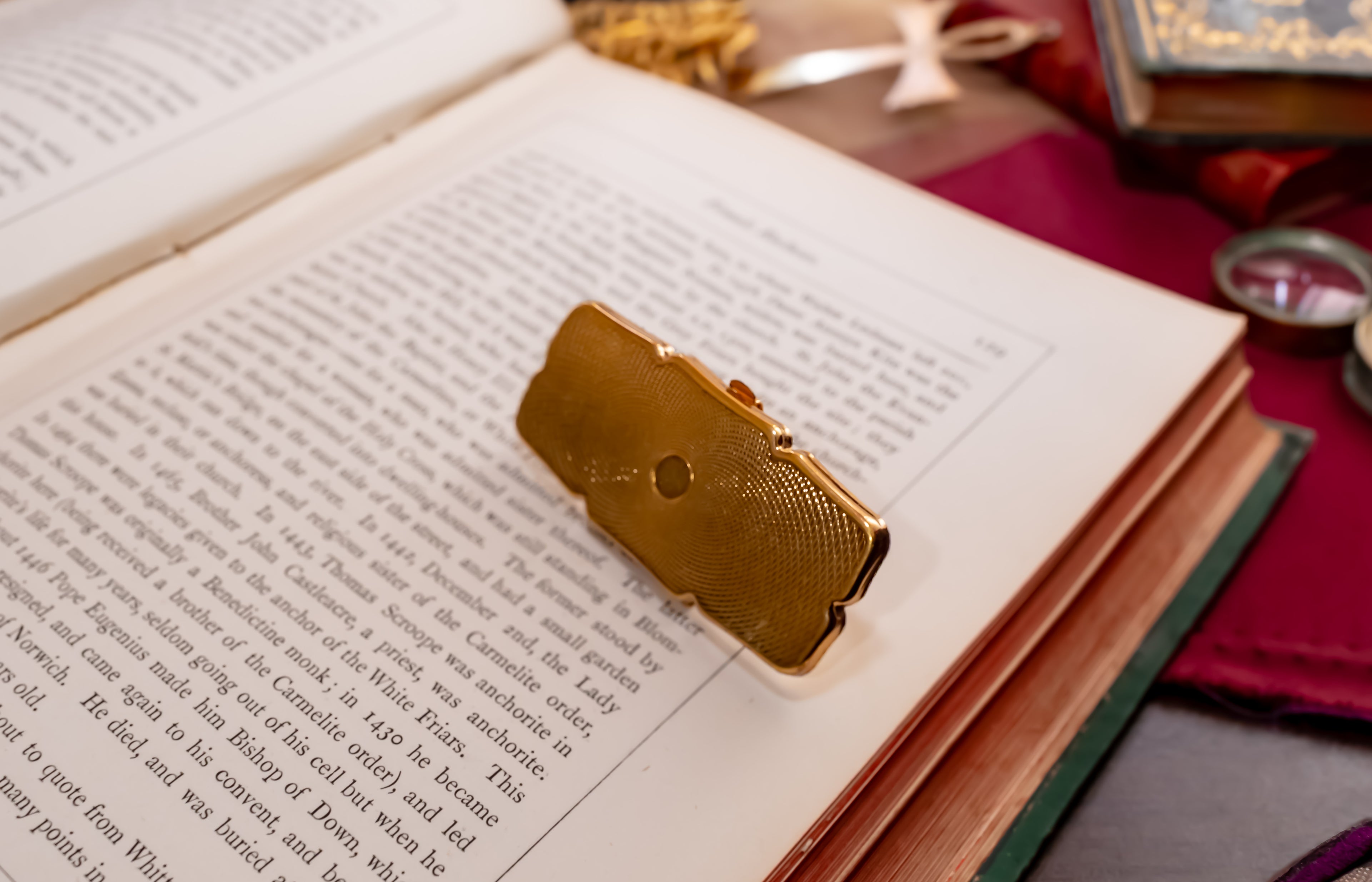 A small rectangular gold-toned Stratton lipstick holder with a textured surface and a circular cutout, resting on the pages of an open book surrounded by vintage-style props.
