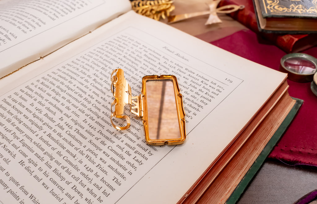 A gold-toned stratton lipstick holder with an open rectangular case rests on the pages of an open book, surrounded by vintage books and a red cloth in a styled tabletop setting.