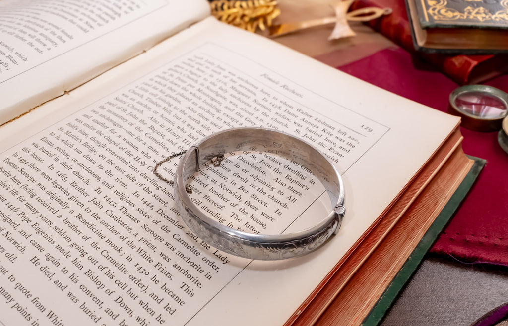 A silver-toned metal cuff bracelet with engraved floral patterns and a small hanging tag, displayed on the open pages of an antique book.