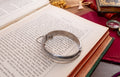 A silver-toned metal cuff bracelet with engraved floral patterns and a small hanging tag, displayed on the open pages of an antique book.