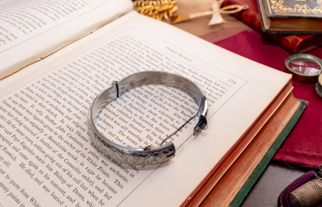 A silver-toned metal cuff bracelet with engraved floral details and a small chain extension, resting on the pages of an open book surrounded by vintage study props.