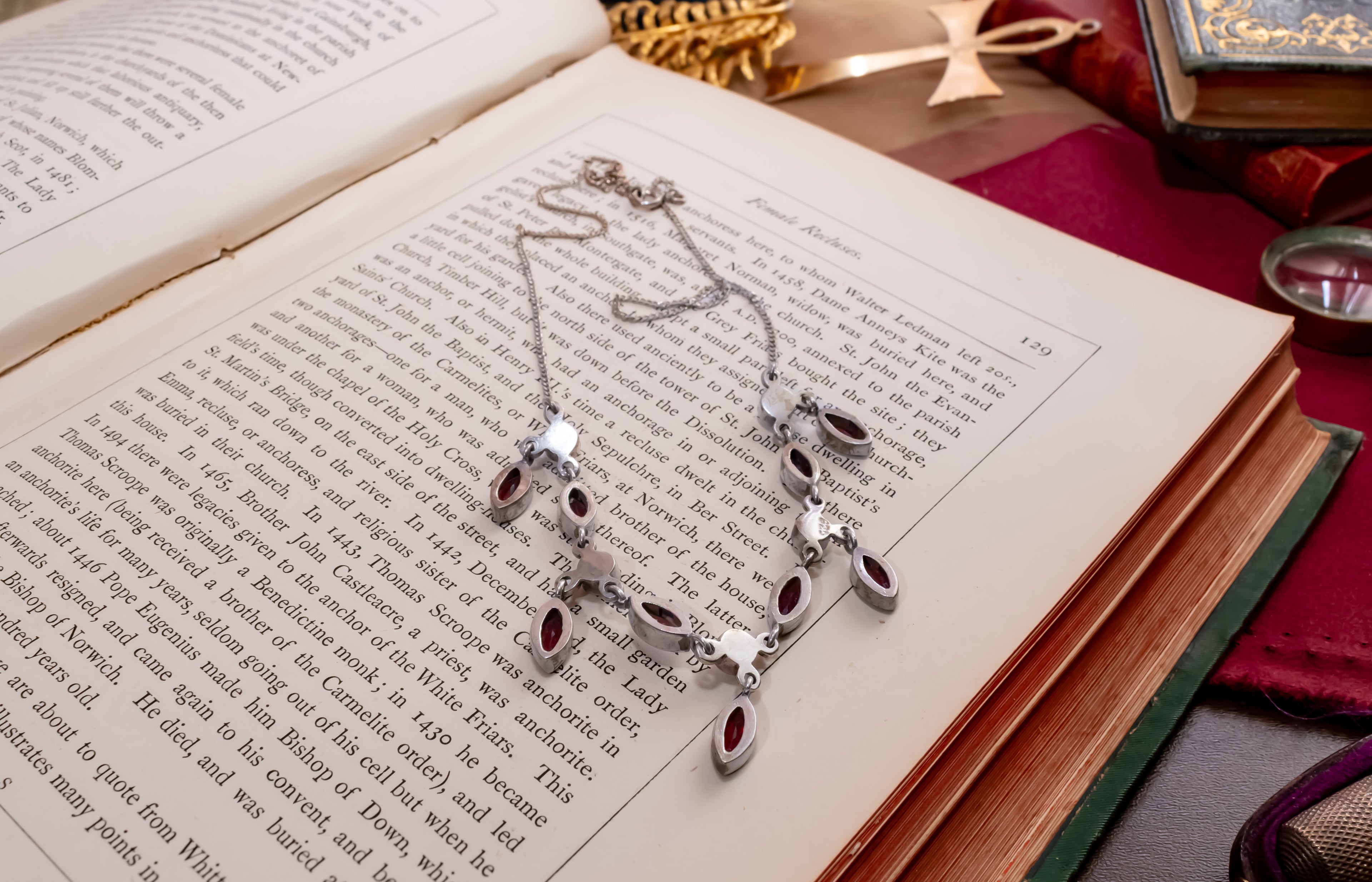 A silver-toned pendant necklace with multiple dangling oval charms, each set with a dark red stone, displayed on the pages of an open book. The scene is styled with vintage props including old books, a gold compass, and a red cloth.
