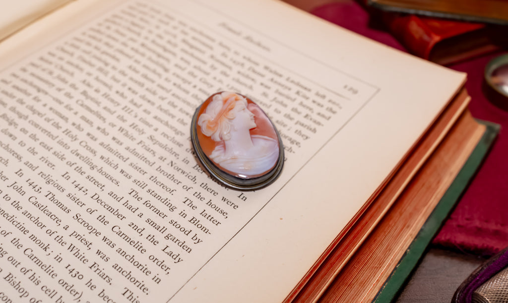 A vintage-style oval cameo brooch with a white and peach portrait on a dark metal setting, resting on the pages of an open antique book.