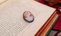 A vintage-style oval cameo brooch with a white and peach portrait on a dark metal setting, resting on the pages of an open antique book.