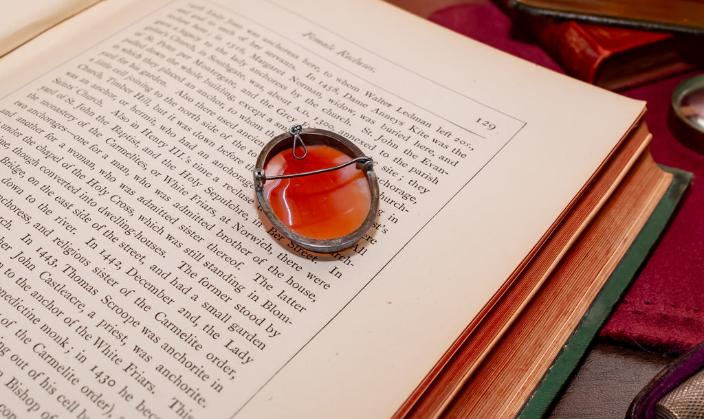 A round amber color cameo with a silver metal frame and pin, resting on an open vintage book with printed text.