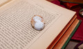 A white carved cameo-style pendant with a gold-toned setting rests on an open vintage book page, surrounded by old books and a red fabric surface.