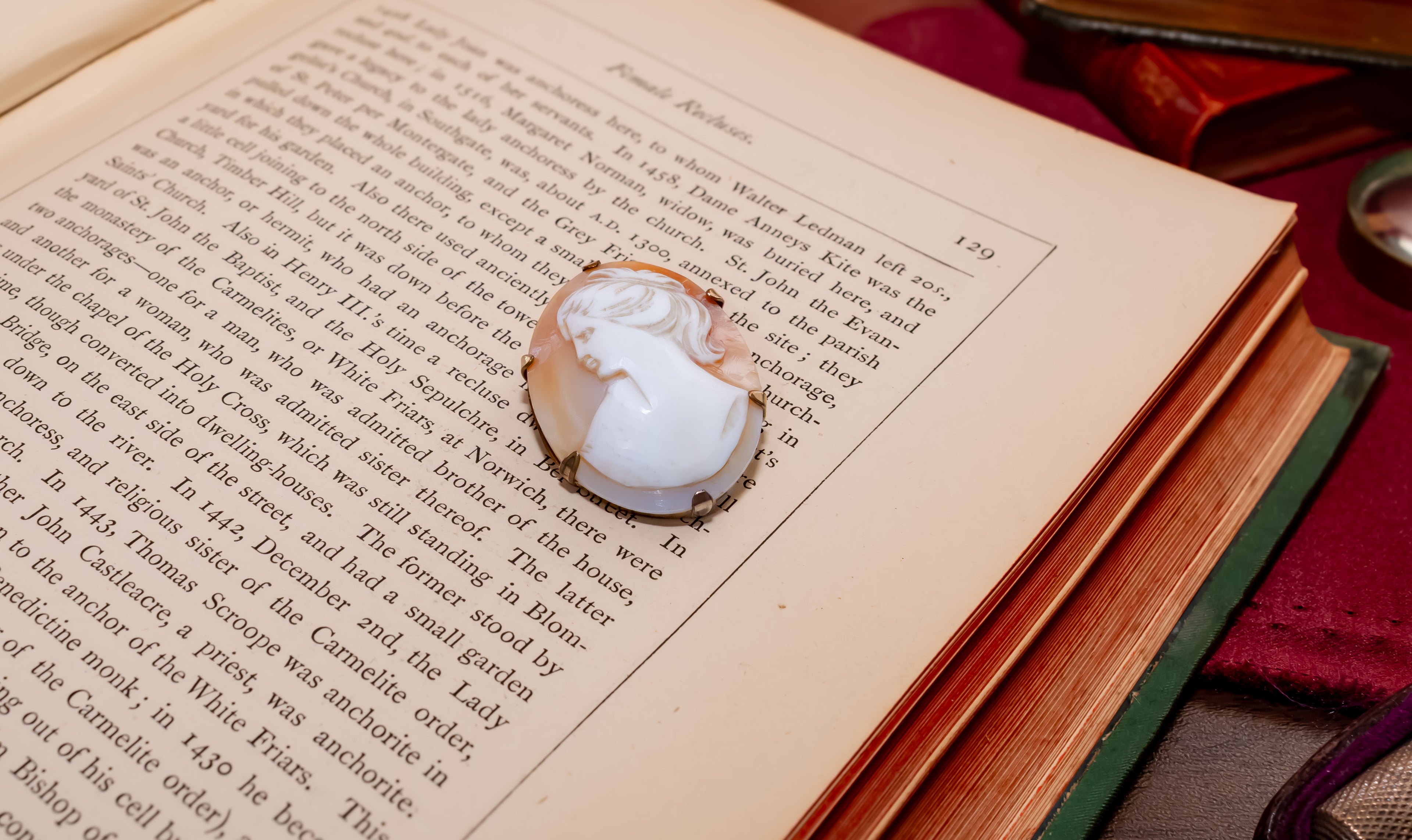 A white carved cameo-style pendant with a gold-toned setting rests on an open vintage book page, surrounded by old books and a red fabric surface.