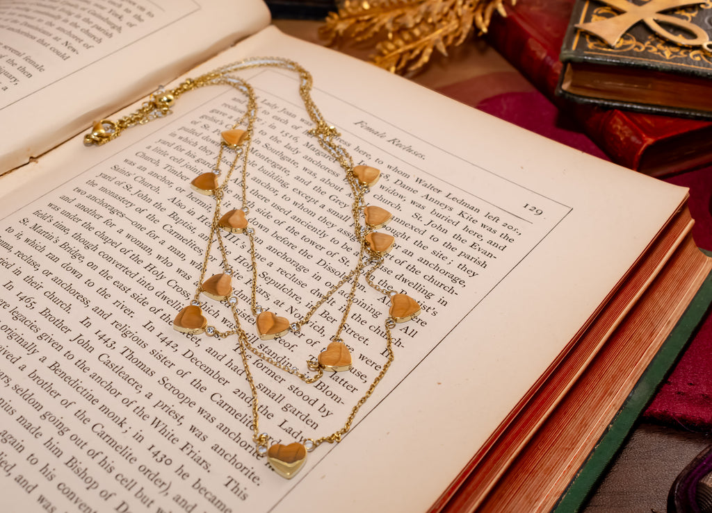 A gold-toned layered necklace with multiple heart-shaped charms draped over an open book, surrounded by vintage books and a gold decorative object.